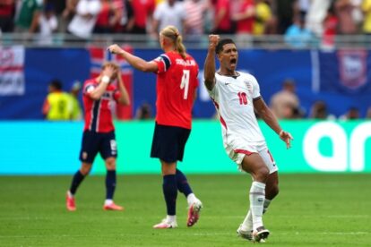 England forward (No.10) celebrates a goal with a raised fist as teammates in red react in the background on a soccer field in front of cheering fans.