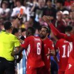 Redjersey soccer players celebrate surrounding the referee after a decision with fans in the stands in the background