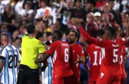 Red‑jersey soccer players celebrate, surrounding the referee after a decision, with fans in the stands in the background.