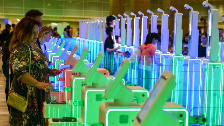 People line up at a row of colorful self service kiosks with blue green and red lighting in a spacious exhibition hall