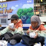 Two kids in hoodies sit on a blue floor painting white paper cups at a craft station during kai mini fair