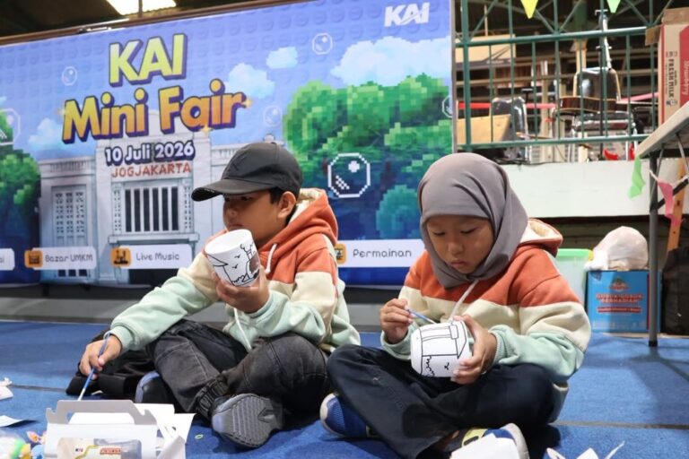 Two kids in hoodies sit on a blue floor, painting white paper cups at a craft station during KAI Mini Fair.