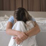 Young woman sits on the floor by a bed arms wrapped around her knees with her head resting on them looking distressed