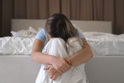 Young woman sits on the floor by a bed, arms wrapped around her knees with her head resting on them, looking distressed.