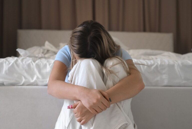 Young woman sits on the floor by a bed, arms wrapped around her knees with her head resting on them, looking distressed.