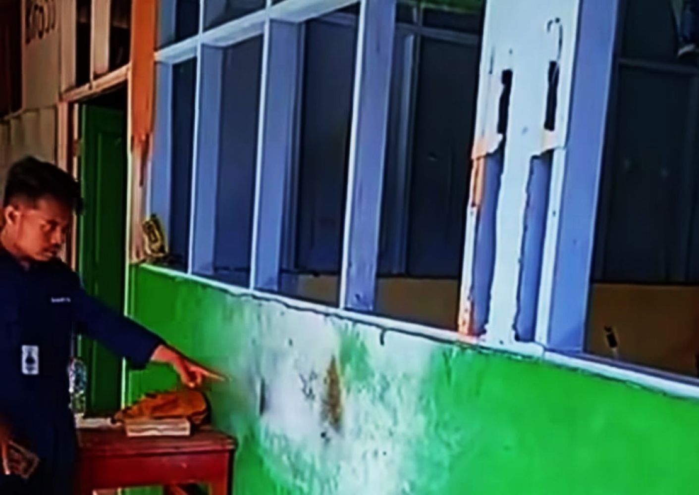 Man in dark jacket pointing at a green painted wall inside a room with a small table and bread on it to his left