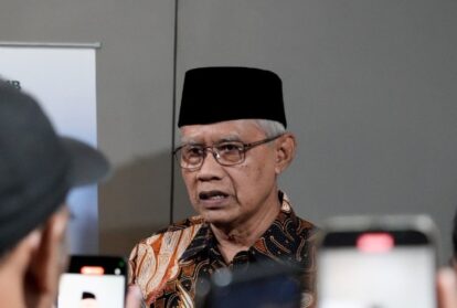Senior indonesian man in batik shirt and black peci hat speaks to reporters smartphones raised around him