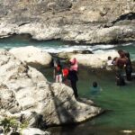 Group of people wading in a turquoise river among large gray rocks on a sunny day