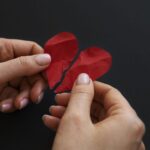 Two hands pull apart two red textured paper hearts on a dark background revealing a split heart shape for a craft project