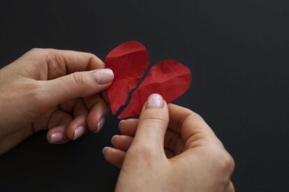 Two hands pull apart two red textured paper hearts on a dark background revealing a split heart shape for a craft project