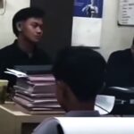 Two people sit at a desk in an office reviewing stacked papers and documents