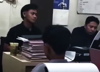 Two people sit at a desk in an office, reviewing stacked papers and documents.