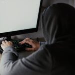 Person in a gray hoodie types on a keyboard in front of a large monitor with a blank pale screen