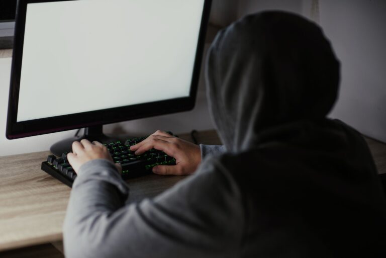Person in a gray hoodie types on a keyboard in front of a large monitor with a blank pale screen.