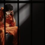Prisoner in an orange jumpsuit sits on a bench in a dim jail cell head in hands behind bars expressing despair