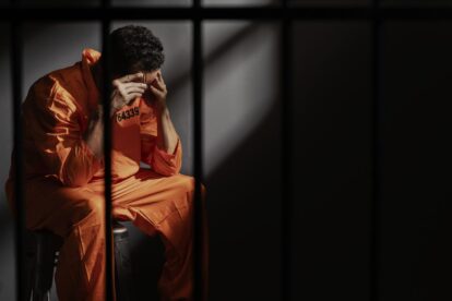 Prisoner in an orange jumpsuit sits on a bench in a dim jail cell head in hands behind bars expressing despair