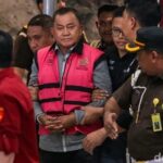 Older man in a pink vest being escorted by uniformed officers through a crowd