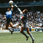 Two goalkeepers leap for the ball during a match one in a blue jersey and the other in gray with a crowded stadium in the background
