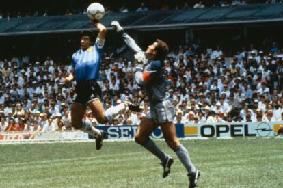 Two goalkeepers leap for the ball during a match one in a blue jersey and the other in gray with a crowded stadium in the background