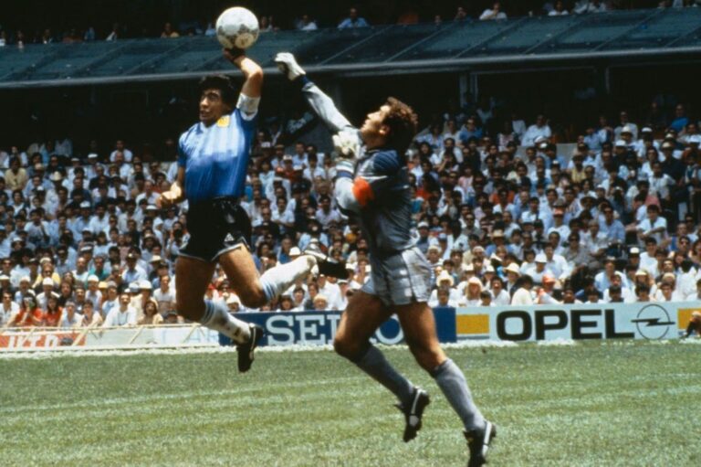 Two goalkeepers leap for the ball during a match, one in a blue jersey and the other in gray, with a crowded stadium in the background.
