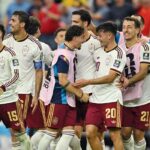 Group of soccer players in white jerseys and maroon shorts celebrate together on the field smiling and embracing each other