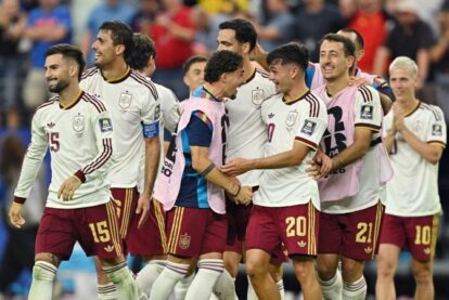 Group of soccer players in white jerseys and maroon shorts celebrate together on the field, smiling and embracing each other.