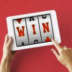 Hands hold a white tablet displaying a three reel slot machine with the word win on a red background