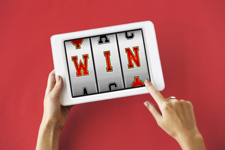 Hands hold a white tablet displaying a three-reel slot machine with the word WIN on a red background.