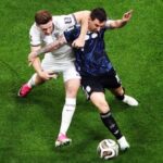 Two soccer players fight for the ball during a match one in white with pink cleats and the other in dark blue on a green pitch