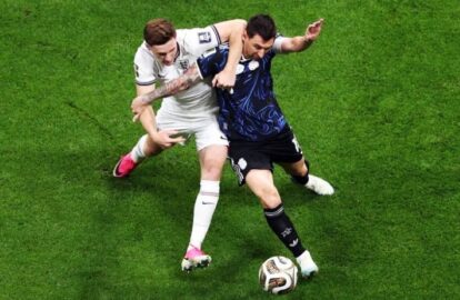 Two soccer players fight for the ball during a match, one in white with pink cleats and the other in dark blue, on a green pitch.
