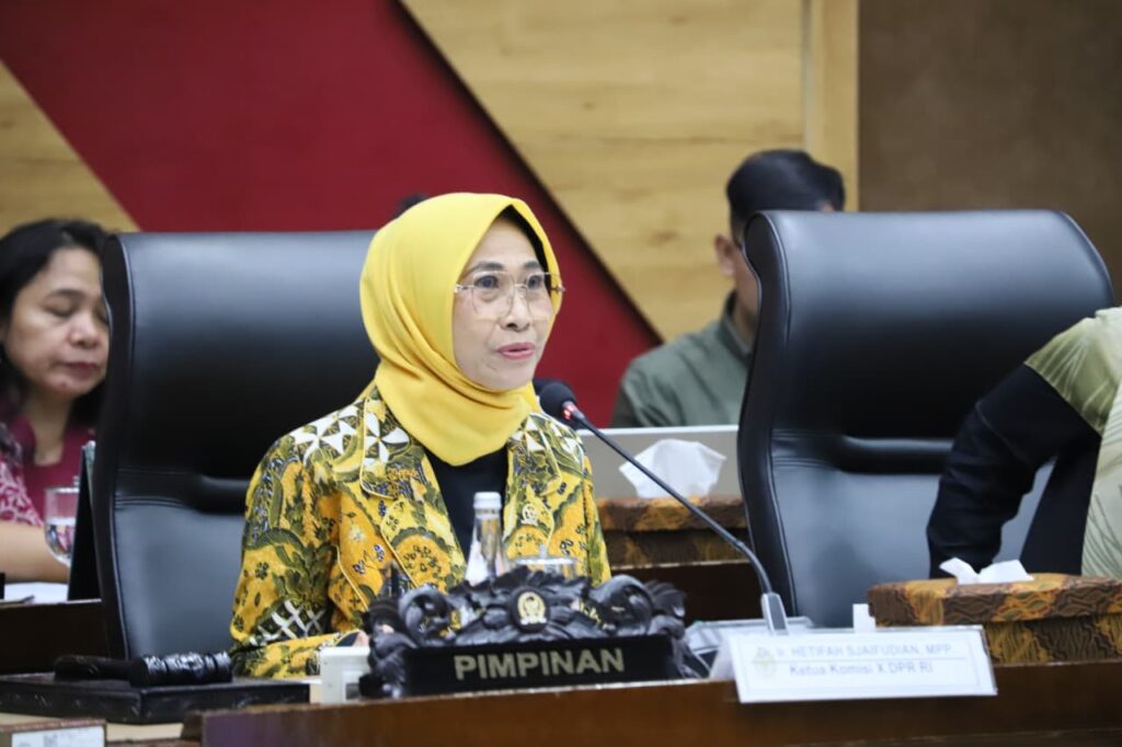 1781683843d1c93e51c27125a7394e catrawarta Woman wearing a yellow hijab and patterned jacket speaks into a microphone at a formal meeting with a'PIMPINAN' sign on the desk in front of her.