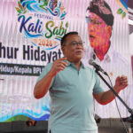 Man in a light blue polo shirt speaking into a microphone on a stage with a colorful festival banner in the background