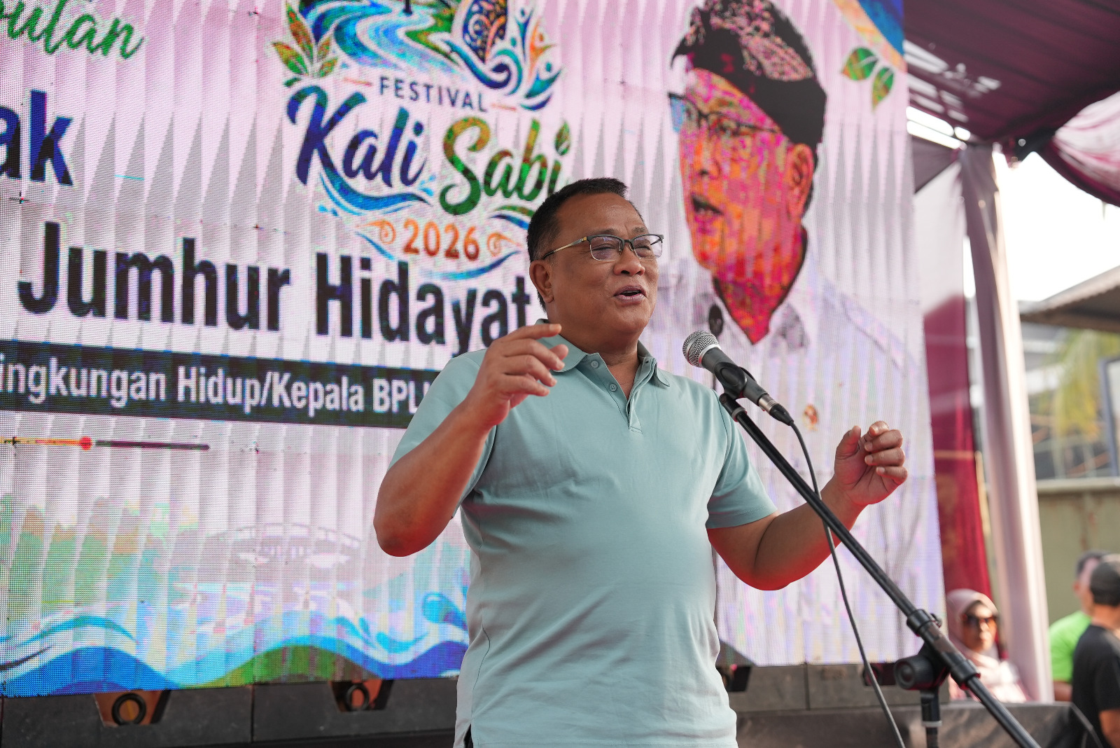 Menteri lingkungan hidupkepala badan pengendalian lingkungan hidup catrawarta Man in a light blue polo shirt speaking into a microphone on a stage with a colorful festival banner in the background
