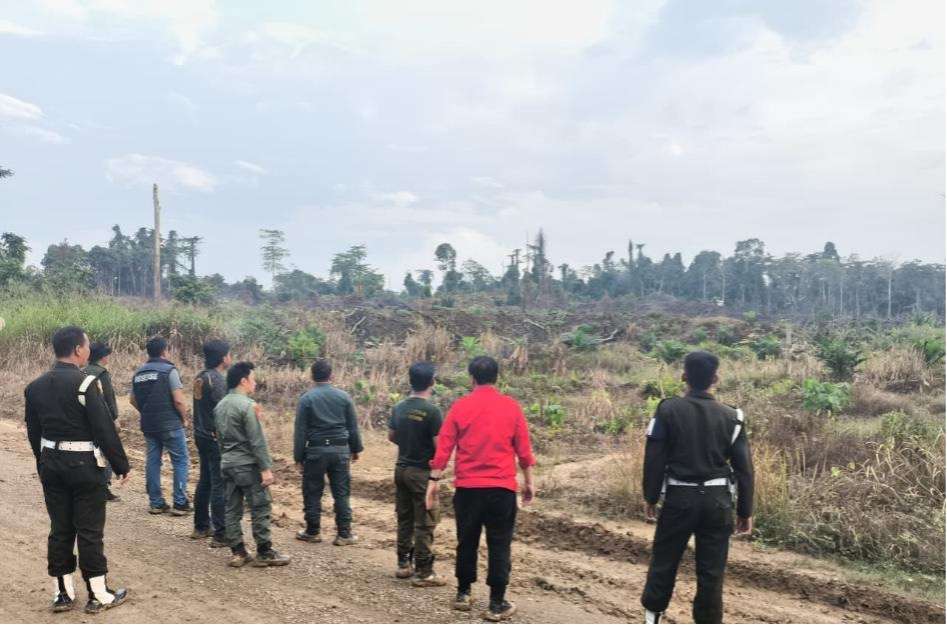 Group of uniformed individuals standing along a dirt road surveying a recently cleared barren field under a cloudy sky