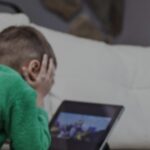 Child in a green shirt lies on a sofa head in hands looking at a tablet screen