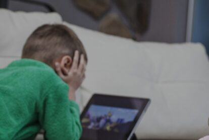 Child in a green shirt lies on a sofa, head in hands, looking at a tablet screen.