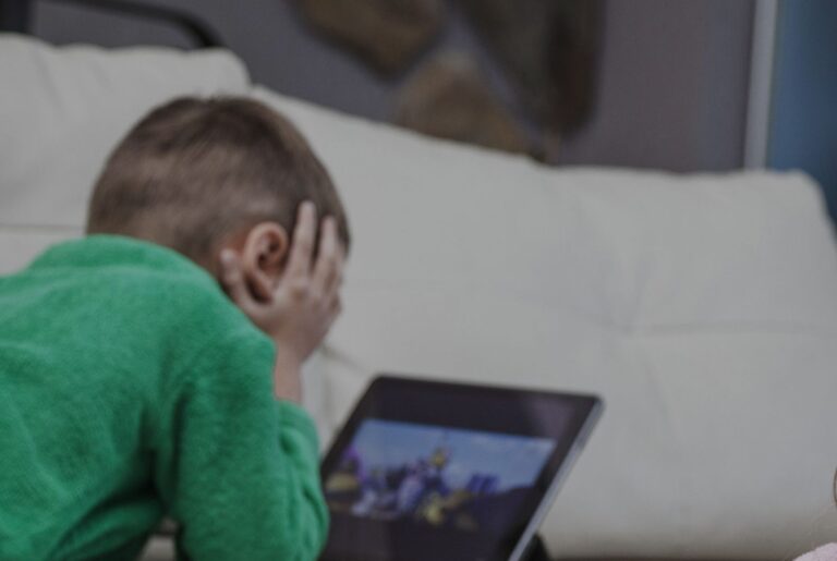 Child in a green shirt lies on a sofa, head in hands, looking at a tablet screen.