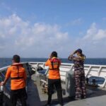 Four crew members on a patrol boat deck scan the sea two wear orange basarnas uniforms one in camouflage all looking outward with binoculars or hands to their eyes