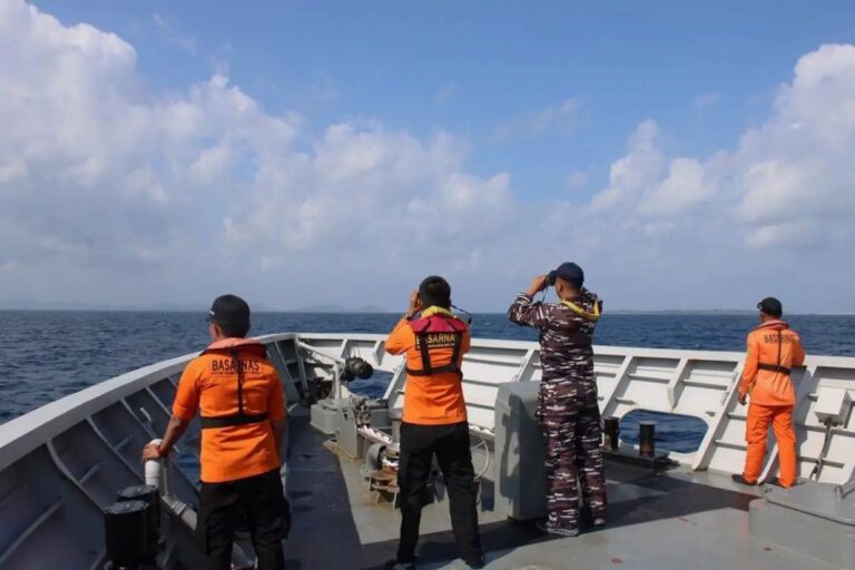 Four crew members on a patrol boat deck scan the sea; two wear orange BASARNAS uniforms, one in camouflage, all looking outward with binoculars or hands to their eyes.
