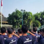 Group of people in matching navy shirts standing together outdoors saluting toward a flagpole with an indonesian flag in a sunny park setting