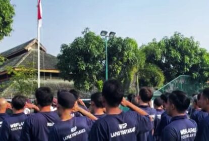 Group of people in matching navy shirts standing together outdoors, saluting toward a flagpole with an Indonesian flag in a sunny park setting.
