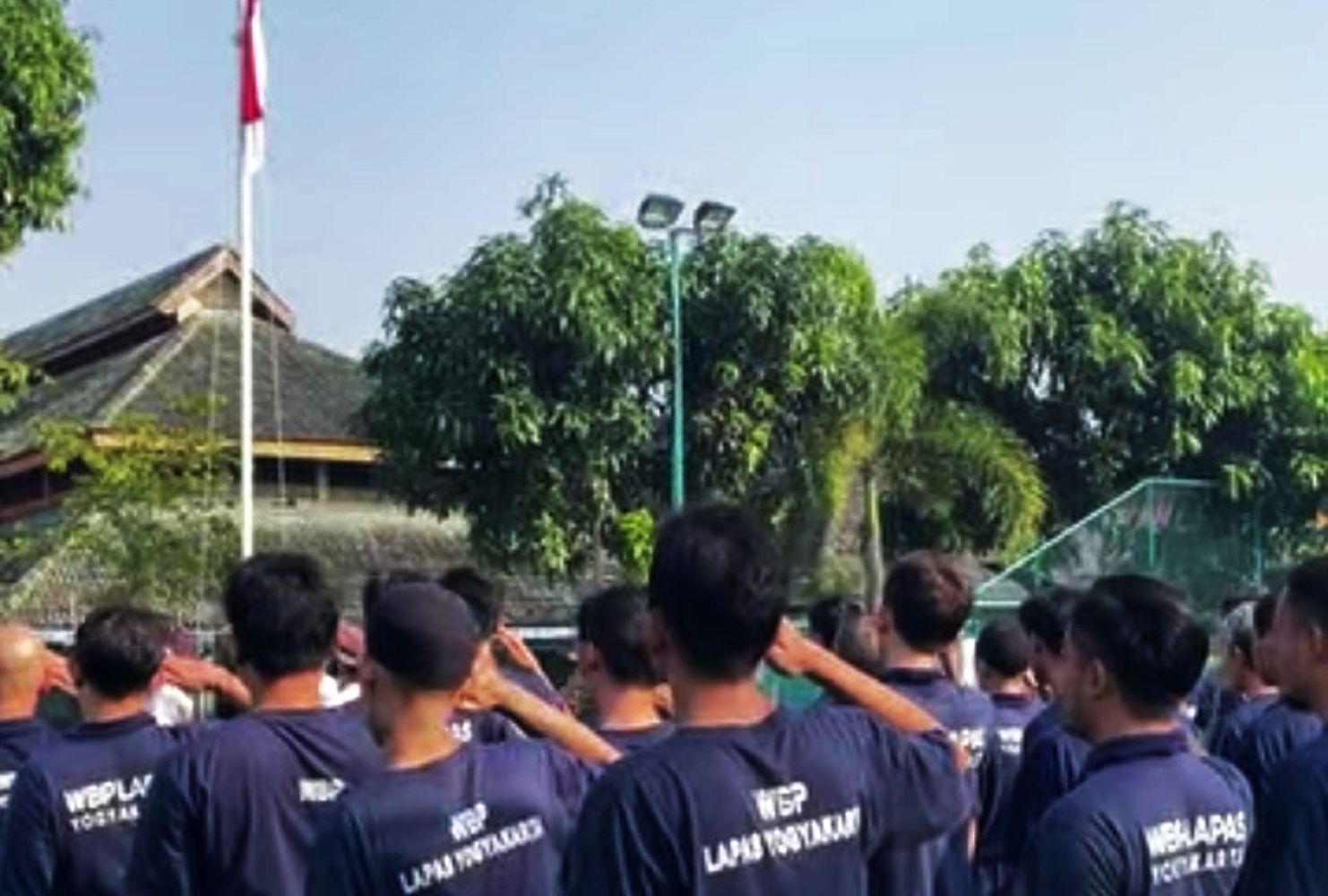 Group of people in matching navy shirts standing together outdoors saluting toward a flagpole with an indonesian flag in a sunny park setting