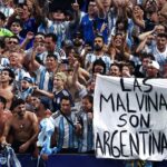 Crowd of argentine soccer fans in striped shirts cheer with raised arms holding a banner that reads las malvinas son argentinas