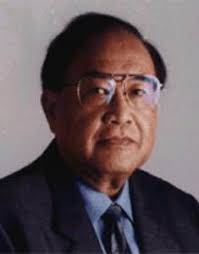 Professional portrait of a middle aged man in a dark suit and blue shirt wearing glasses facing the camera with a neutral expression