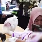 Woman wearing a pink hijab sits beside a hospital bed looking down at a patient with an iv stand nearby In a clinical setting with medical equipment and bags on the table behind her