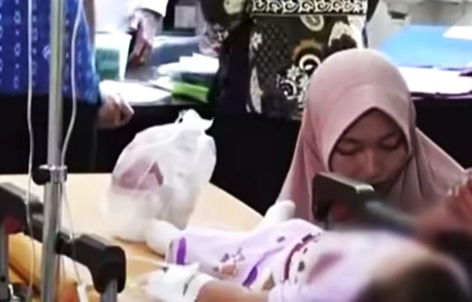 Woman wearing a pink hijab sits beside a hospital bed looking down at a patient with an iv stand nearby In a clinical setting with medical equipment and bags on the table behind her