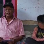 Two people sit side by side on a bench outside an older man in a pink plaid shirt and black cap on the left and a younger person with a large arm scar on the right