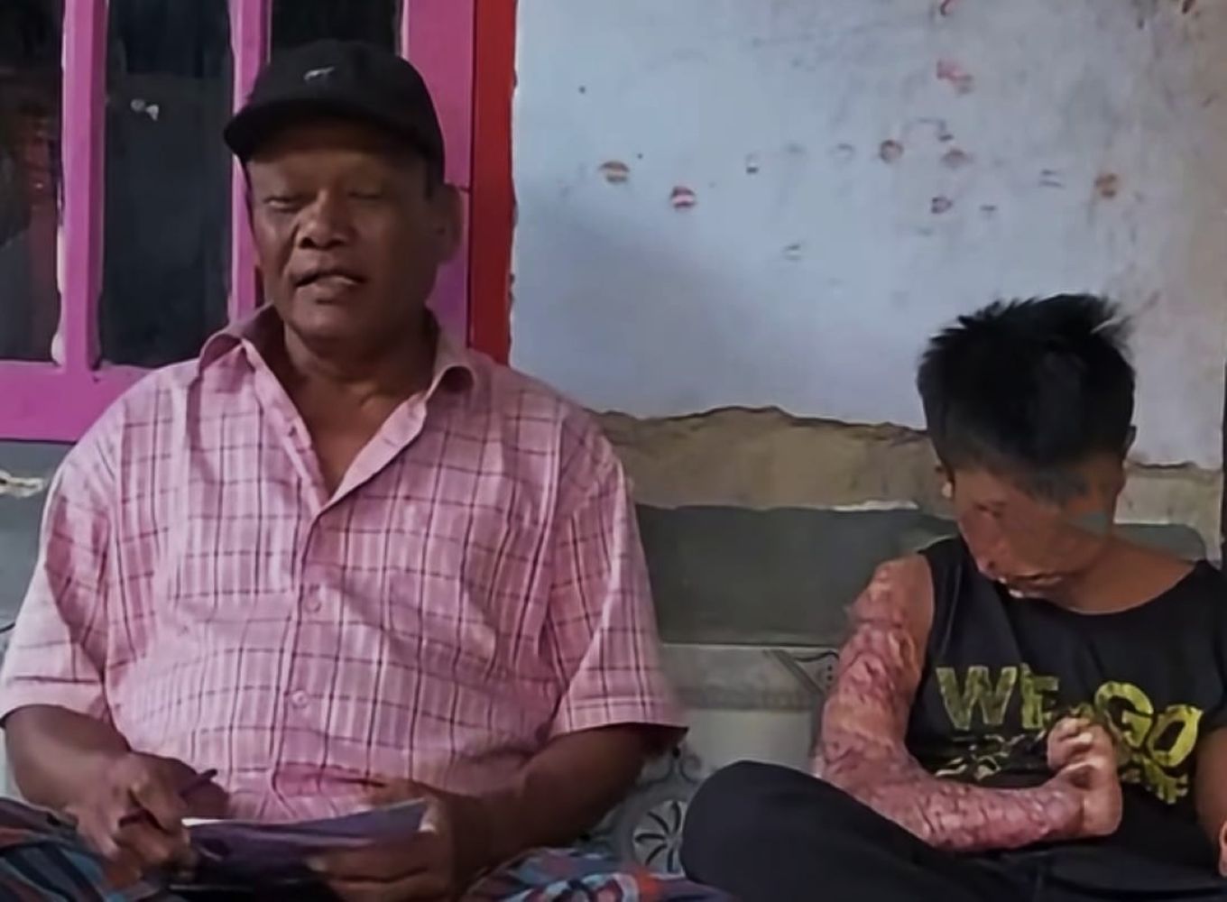 Two people sit side by side on a bench outside an older man in a pink plaid shirt and black cap on the left and a younger person with a large arm scar on the right