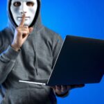 Person in a gray hoodie wearing a white mask holds a laptop and shushes with a finger to lips suggesting a hacker or secretive activity