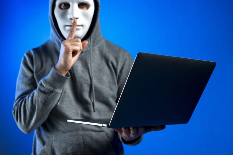 Person in a gray hoodie wearing a white mask holds a laptop and shushes with a finger to lips, suggesting a hacker or secretive activity